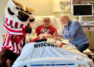 Bucky Badger giving a thumbs up with patient in memory care suites bed